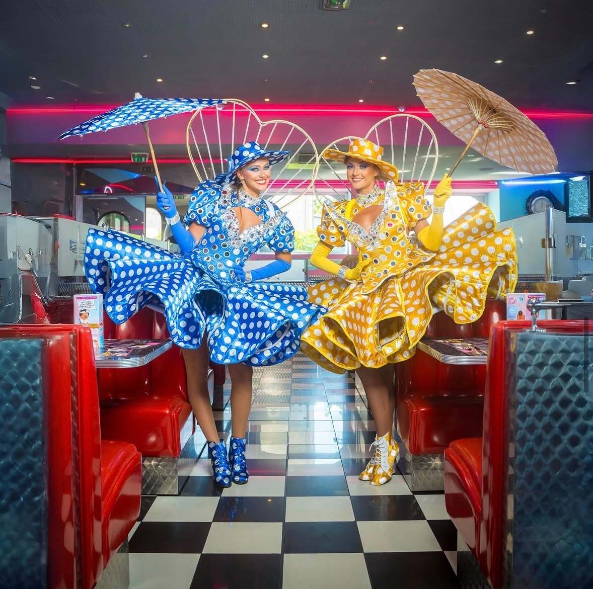 Deux artistes en robes à pois bleue et jaune, ombrelles ouvertes, entre les banquettes d'un diner