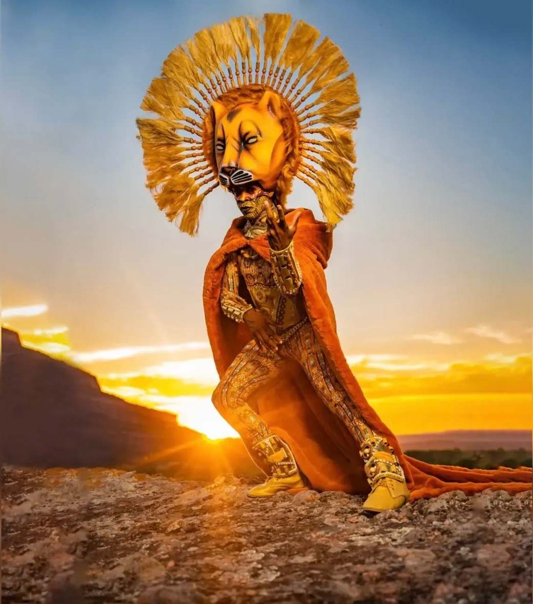Artiste en costume de lion à la crinière rayonnante, agenouillé face au soleil couchant